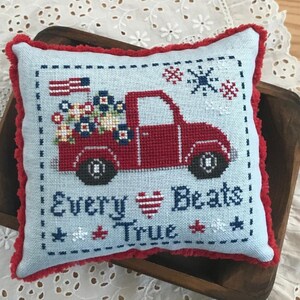 May include: A red truck with a blue and white American flag and flowers in the back. The truck is on a blue and white pillow with the text "Every Heart Beats True".
