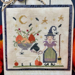 May include: Cross-stitch design titled "Boo! Ewe" featuring a witch, sheep, pumpkins, and a crescent moon. The design is framed and mounted on a decorative wooden stand. The text "The Proper Stitcher" is visible.