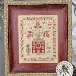 May include: Framed cross-stitch sampler in a red and gold ornate frame. The sampler depicts a house, flowers, birds, and the text "ABC ~ 123 SAMPLER". The design is by Brenda Gervais.