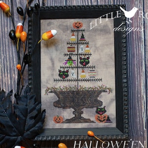 May include: A cross-stitch pattern of a Halloween feather tree with a black cauldron, pumpkins, ghosts, and black cats. The pattern is framed in a black frame and the text "HALLOWEEN FEATHER TREE" is below the frame. The text "LITTLE ROBIN designs" is in the upper right corner of the image.