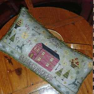 May include: A rectangular, embroidered pillow with a Christmas theme. The pillow features a red house, Christmas trees, reindeer, and snowflakes on a light blue background. The text "HOME FOR CHRISTMAS SAMPLER PILLOW" is at the top. The pillow is on a wooden table.