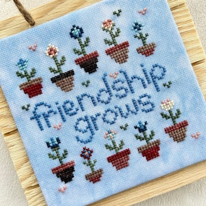 May include: A blue cross-stitch embroidery with a wooden frame. The embroidery features a variety of colorful flowers in pots and the text "Friendship grows".