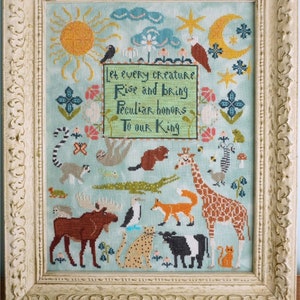 May include: A framed cross-stitch embroidery featuring a variety of animals, including a giraffe, a moose, a fox, a cat, and a beaver. The embroidery is set against a light blue background with a green border. The text "Let every creature rise and bring peculiar honors to our King" is stitched in the center of the embroidery.