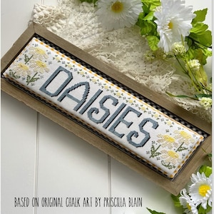 May include: A framed cross-stitch design featuring the word "DAISIES" in blue lettering. The design is surrounded by a yellow and white checked border. The frame is brown wood with a white background. The image includes the text "Stitching with the Housewives" and "May Word Swap".