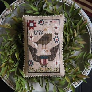 May include: A beige, rectangular decorative pillow with a cross-stitch design featuring an eagle, stars, and the year 1776. The pillow is surrounded by a white beaded tray with greenery. The text "FREEDOM FLOCK" is displayed below the pillow.
