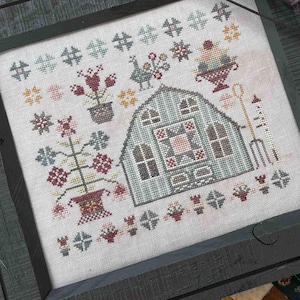 May include: A framed cross-stitch sampler featuring a greenhouse design with flowers and a garden tool. The frame is dark green, and the sampler is set on a patchwork quilt. A light blue bird figurine and dried lavender sprigs are nearby.