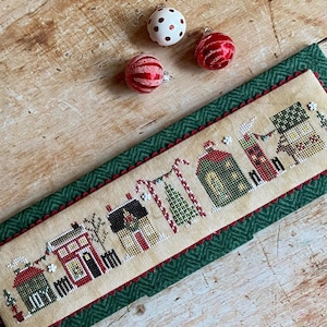 May include: A cross-stitch sampler featuring a row of houses decorated for Christmas with candy canes, lights, and a Christmas tree. The sampler is framed with a green fabric with a red border.