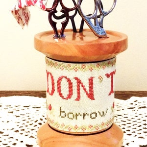 May include: A wooden spool with a white fabric cover that says "Don't Borrow" in red letters. The spool is holding three pairs of scissors, two with red handles and one with silver handles.