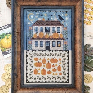 May include: A framed cross-stitch pattern featuring a white house with a black cat and a crow on the roof. The house is surrounded by pumpkins and vines. The background is a blue sky with stars and spiders.