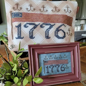 May include: A framed cross-stitch featuring the American flag and the year "1776" in navy blue. The frame is a muted red-brown color. A matching pillow with the same design is in the background. The design includes anchors.