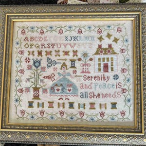 May include: Framed cross-stitch sampler with a gold-colored ornate frame. The sampler features a house, floral elements, and the text "Serenity is all She needs." A pair of decorative scissors rests below the frame.