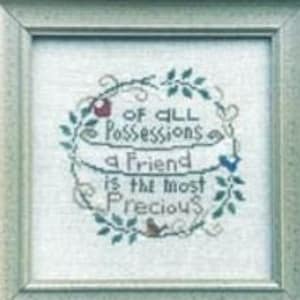 May include: A cross-stitch pattern featuring a wreath of green leaves and a quote that reads "Of all possessions a friend is the most precious". The pattern is framed in a light wood frame and the title "Precious Friend" is written in black text on a wooden background.