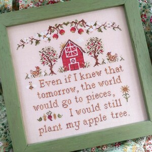 May include: A cross-stitch embroidery framed in a green wood frame. The embroidery depicts a red barn with a white door and windows, two apple trees, and a bee. The text reads: "Even if I knew that tomorrow, the world would go to pieces, I would still plant my apple tree."