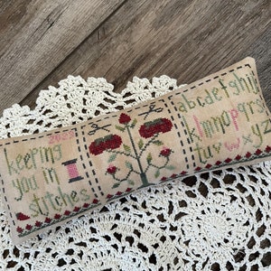 May include: A beige pillow with cross-stitch embroidery. The pillow features a red and green floral design with the text "Keeping you in stitches" and the alphabet in a separate section. The pillow is on a white crocheted doily.