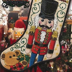 May include: A white and beige Christmas stocking with a Nutcracker soldier design. The stocking has a red and blue uniform, a black hat, and a white beard. The stocking is decorated with gold stars and green vines. The name "Natalia" is embroidered on the stocking.