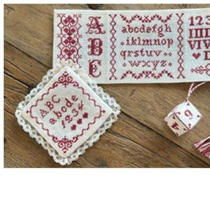 May include: A set of five cross-stitch embroidery pieces with a white background and red stitching. The pieces include an alphabet, numbers, a crown, a dice, and a small square with the numbers 2025.