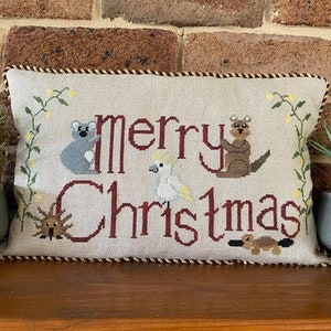May include: A beige pillow with a red embroidered border and the words "Merry Christmas" in red. The pillow features embroidered images of Australian animals, including a koala, a kookaburra, a cockatoo, an echidna, and a platypus.
