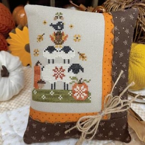 May include: A decorative pillow with a cross-stitch design featuring sheep, a bird, and pumpkins. The pillow has a beige background with orange and brown accents. The text "AUTUMN EWE" and "PRIMROSE COTTAGE" are visible.