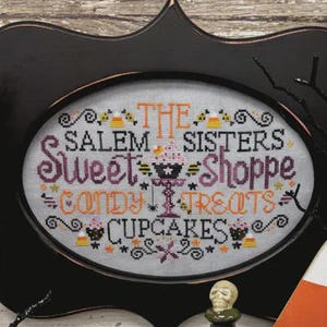 May include: A Halloween-themed display featuring a framed cross-stitch with "Salem Sisters Sweet Shoppe" text, alongside decorative items like a ghost, pumpkin, and bottles. The color palette includes black, orange, and white.