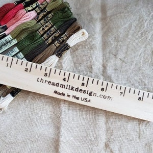 May include: A collection of embroidery floss in various colors, including red, pink, green, and white, is arranged next to a wooden ruler. The ruler, marked in inches, has the text "threadmilkdesign.com" and "Made in the USA" printed on it.