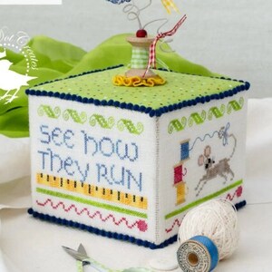 May include: A cross-stitched box with the text "See How They Run" and a mouse design. The box has a green polka-dot top and blue trim. A spool of thread and scissors are in the foreground.