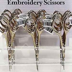 May include: A display stand with gold embroidery scissors. The stand is made of clear acrylic and has two tiers. The scissors are arranged in rows on the stand. Each scissor has a small tag that says "Made in China".