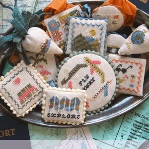 May include: A collection of small, hand-stitched cross-stitch ornaments with travel-themed designs. The ornaments are displayed on a silver tray with a passport and map in the background. The text "FESTIVE LITTLE FOBS TRAVEL EDITION" is displayed at the bottom of the image.