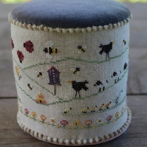 May include: A cylindrical pincushion with a dark gray velvet top and a beige fabric body. The body is embroidered with a whimsical design of bees, birds, flowers, and a birdhouse. The base and top edge are trimmed with a cream-colored pom-pom trim.