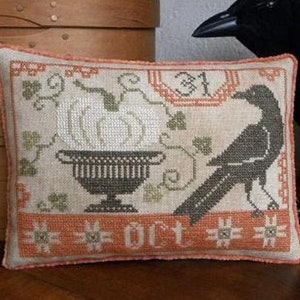 May include: A beige pillow with an embroidered design of a pumpkin in a pot and a black crow. The pillow has an orange border with the text "31 OCT" embroidered on it.