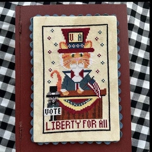 May include: A cross-stitch embroidery of a cat dressed as Uncle Sam, holding a flag and standing behind a podium with the words "Vote" and "Liberty for All".