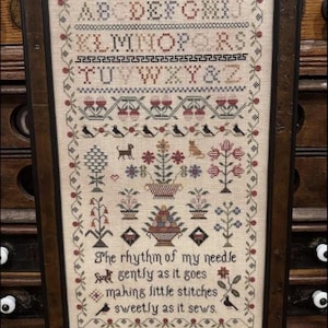 May include: Framed cross-stitch sampler with the text "The Rhythm of My Needle." The design features an alphabet, floral motifs, birds, and decorative borders in shades of red, blue, and green. The sampler is in a dark brown wooden frame.