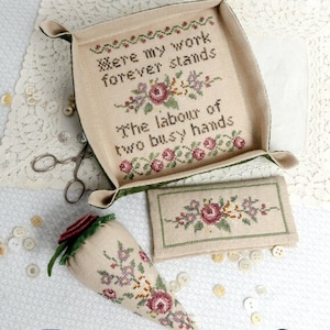 May include: A vintage needlepoint kit with a green and beige tray, a small beige pouch, and a beige pincushion shaped like a carrot. The tray has a floral design and the text "Here my work forever stands The labour of two busy hands".