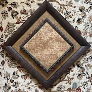 May include: A framed needlepoint sampler with a dark brown textured frame. The sampler features a poem with red and black lettering. The frame is diamond-shaped, with a smaller, square frame inside. The background is a patterned rug.