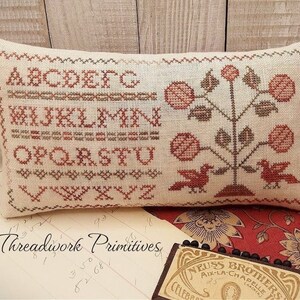 May include: A beige pillow with a cross-stitch design featuring the alphabet in red and green thread. The pillow also has a cross-stitch design of a stylized tree with red flowers and two birds.