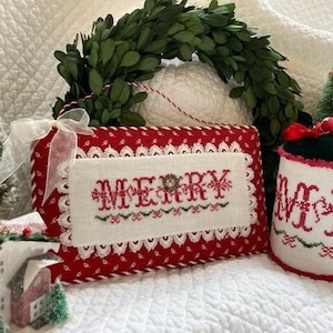 Puede incluir: Una almohada roja y blanca con un borde de encaje blanco y la palabra "Merry" bordada en rojo y verde. La almohada está sobre una superficie blanca.