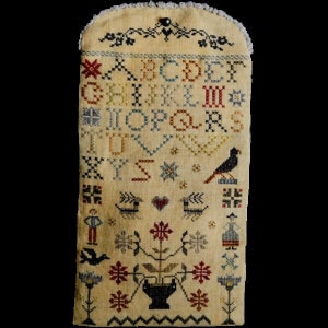 May include: A beige sampler with a cross-stitch design featuring the alphabet, a bird, a heart, a flower, and a small figure. The sampler is framed with a white border and has a button at the top.