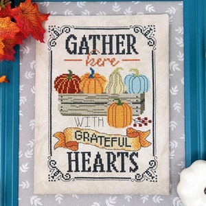 May include: Cross stitch pattern with a teal frame and a white background. The pattern features a basket of colorful pumpkins with the text "Gather here with grateful hearts".