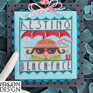 May include: A cross-stitch pattern with a blue background and a girl wearing sunglasses and a hat. The text "Resting Beach Face" is above the girl and the text "ARE YOU BEACH READY?" is below the girl. The pattern is framed in a black and gray frame.