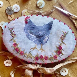 May include: An oval needle book with a cross-stitched blue hen design, surrounded by a floral wreath. The text "Little French Hen Needle Book" and "Stacy Nash Designs" are visible. The image also includes buttons, a thimble, and gold scissors.