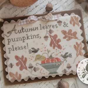 May include: A rectangular cross-stitch sampler featuring the text Autumn leaves & pumpkins, please! with embroidered motifs of oak leaves, acorns, birds, and a bowl of pumpkins. The fabric has scalloped edges and a small beige gingham ribbon bow.