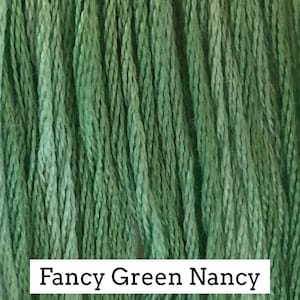 May include: Close-up of a bundle of green embroidery floss. The floss is a variegated green color, with shades of light and dark green. The text "Fancy Green Nancy" is printed on a white label at the bottom of the image.