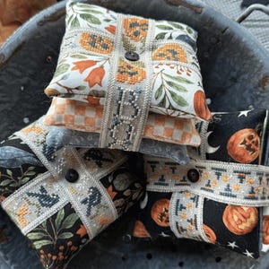 May include: A stack of decorative, square fabric items with button closures, featuring Halloween-themed patterns of pumpkins, bats, and floral designs. The items are arranged in a metal colander with fall leaves and the text "THAT'S A WRAP Spooky Edition".