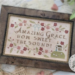 May include: A framed cross-stitch sampler with the words "Amazing Grace, How Sweet the Sound!" in brown thread. The sampler features floral and church motifs in shades of pink, green, and brown. The frame is a distressed brown color.