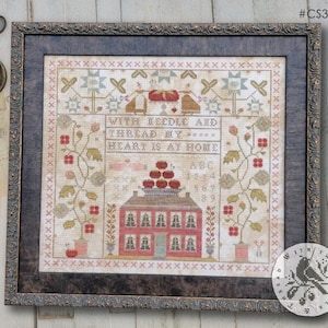May include: Framed cross-stitch sampler with the text "WITH NEEDLE & THREAD, MY HEART IS AT HOME." The design features a red house, floral elements, and a decorative border. The frame is dark with a detailed pattern.