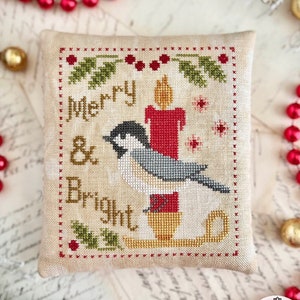 May include: A cross-stitch design on a beige fabric pillow featuring a bird perched on a sled with a lit candle. The design includes the text "Merry & Bright" and a holly sprig.