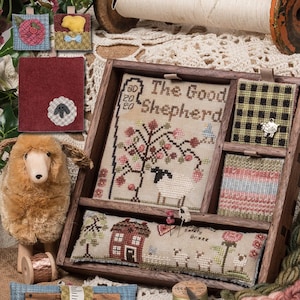 May include: A wooden box with a variety of cross-stitch embroidery pieces inside. The box is labeled "Shepherd's Box" and the embroidery pieces include "The Good Shepherd", a house, and a field of flowers. The box is surrounded by yarn, fabric scraps, and other craft supplies.