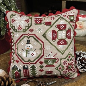 May include: A cross-stitch pillow with a red, green, and white design featuring a snowman, a cup of cocoa, and a snowflake. The pillow is on a wooden tray with pine cones and a red and white string of beads. The text "Cocoa Quaker" and "Primrose Cottage" are visible in the image.