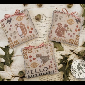 May include: Three small, square, cross-stitch pillows with autumn-themed designs. Each pillow features a different design: a bird, a squirrel, and owls. The pillows are decorated with leaves, berries, and the words "Hello Autumn!"