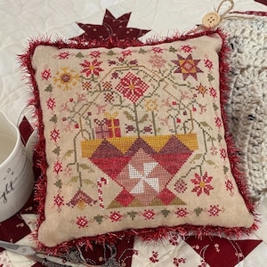 Peut inclure: Un coussin brodé en point de croix rouge et blanc avec un motif de Noël festif. Le coussin est décoré d'une frange rouge, blanche et verte. Le coussin est posé sur une courtepointe rouge et blanche.