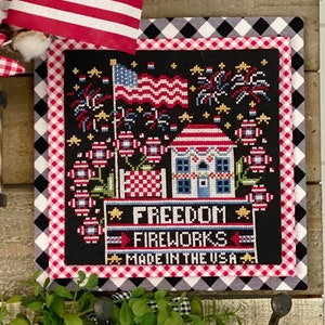 May include: A wooden wall decor with a red, white, and blue American flag, a red and white gingham bow, a glass jar with a scroll inside, a black and white checkered fabric with a cross-stitch design of a house with fireworks and the text "Freedom Fireworks Made in the USA", three hooks with greenery, and a small wooden sign with the text "America the Beautiful".